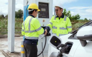 Two technicians shaking hands by a charging station for electric vehicles in bright daylight.